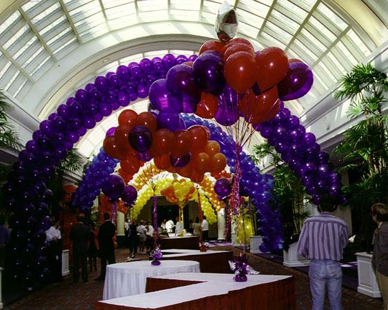This series of packed balloon arches in vivid magenta and citrine yellow colors are placed to decorate a long concourse hall between two event venues forming a visual tunnel focusing attention on the displays placed in the hall.
